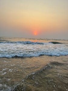 A serene ocean scene at sunset with gentle waves lapping at the shore and a glowing orange sun nearing the horizon, casting a warm reflection on the water.
